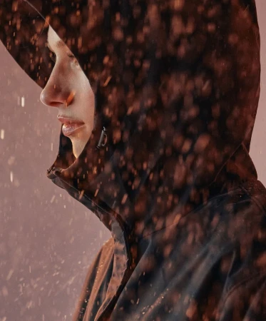 Profile of an adult in a hooded jacket, seen in a close-up shot with an orange-toned color grade and light sprinkling. Fashion and beauty.