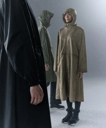 Models standing in falling rain wearing long hooded waterproof raincoats and boots in a studio setting.