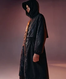 Model in a black hooded raincoat with subtle metallic sheen posing against a warm gradient background