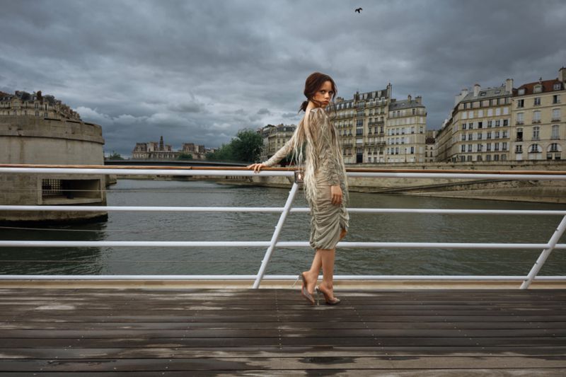 Adult female in an embellished dress stands on a bridge with metal railings and wood floors, and buildings and overcast sky in the background.