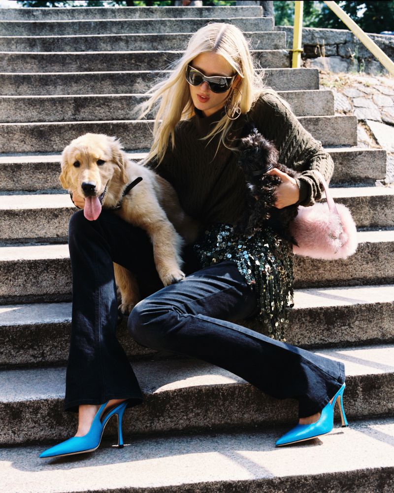 Woman in sunglasses sits on concrete steps with a golden retriever puppy, dressed in dark sweater, jeans, and bright blue high heels.
