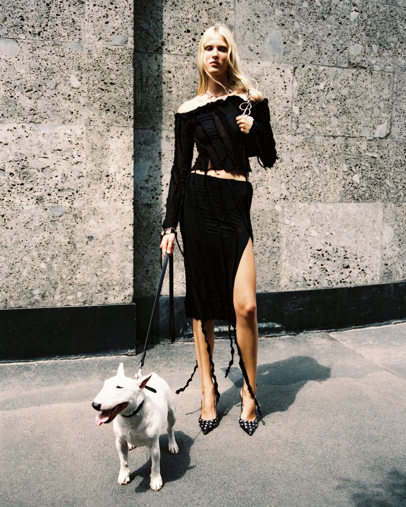 Fashion model walks white bull terrier dog outdoors. Woman in black off-shoulder top and skirt, high heels, stands on sidewalk near wall.
