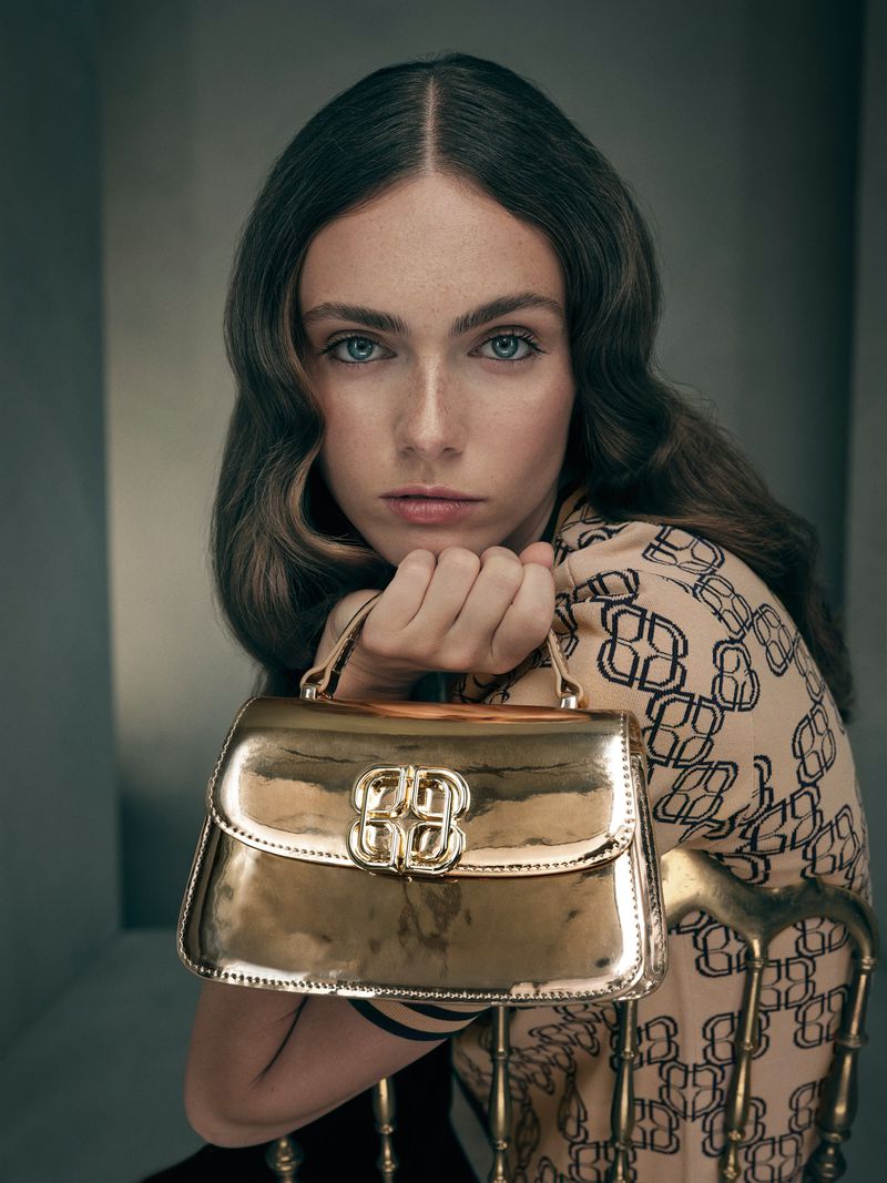 Woman poses indoors with a gold handbag, featuring wavy brunette hair, wearing a patterned top, and sitting on a gold chair.