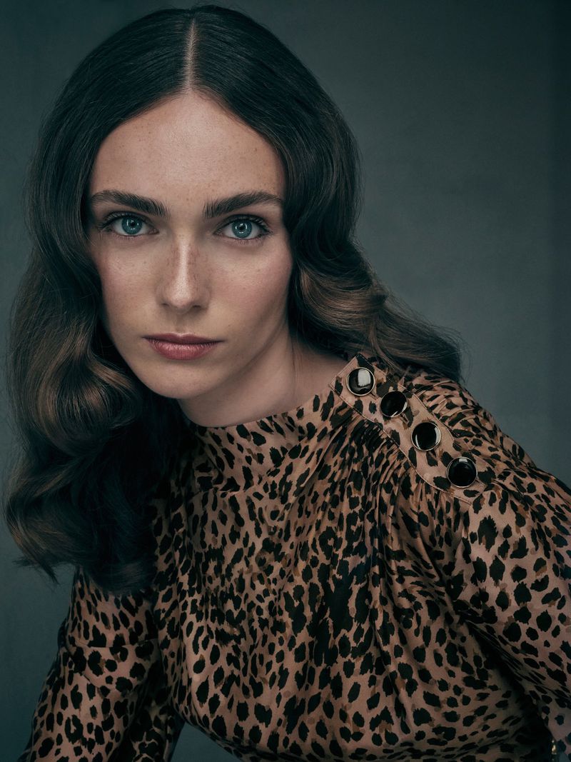 Portrait of a light-skinned adult woman with auburn hair and blue eyes, wearing a leopard-print top against a grey backdrop.