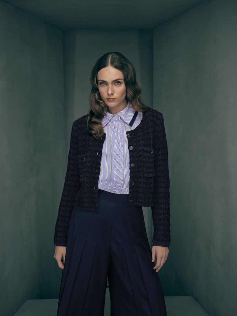 Woman poses indoors wearing a dark jacket, striped collared shirt, and dark pleated culottes, captured in a medium shot.