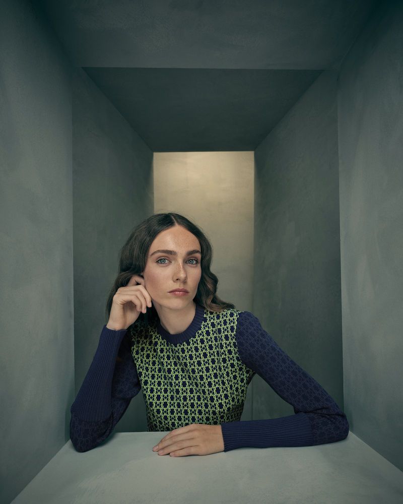 An adult female sits at a white table in a gray corridor wearing a patterned top. The image features a medium shot and eye-level angle.