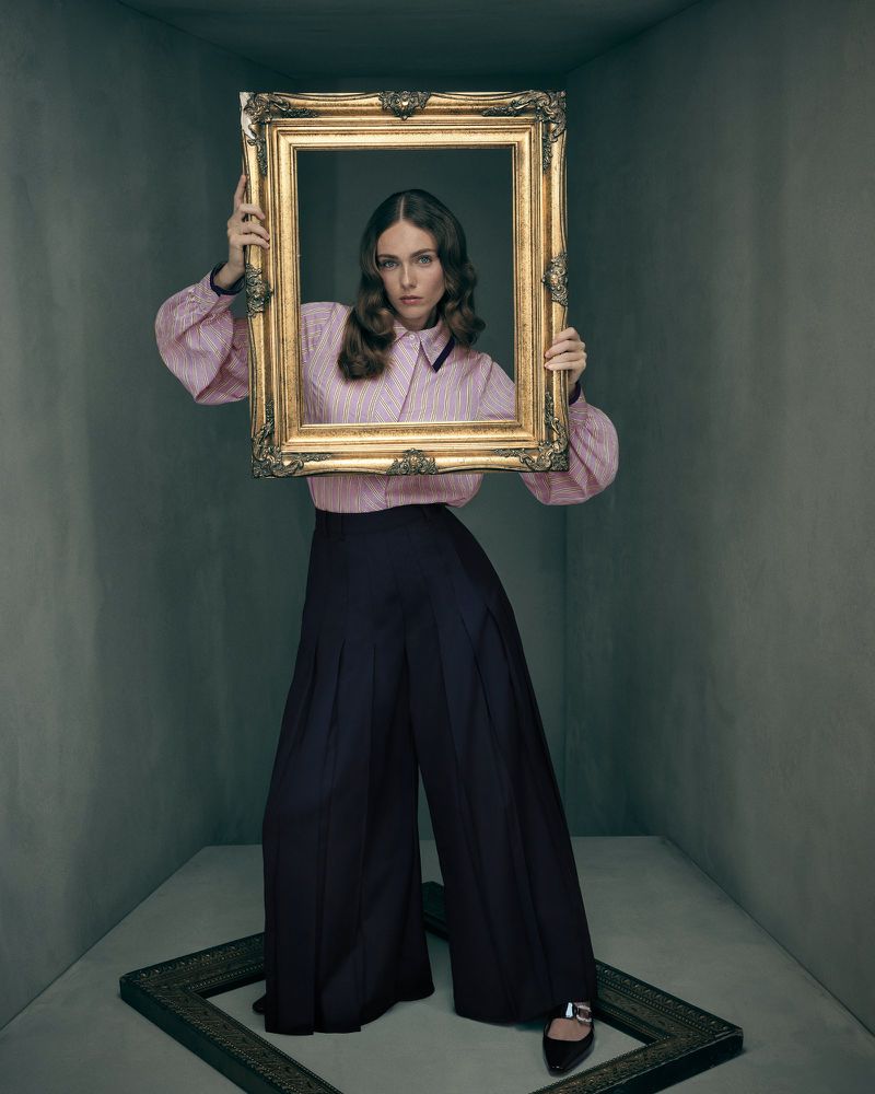 Woman in pink blouse and dark pants holds a gold picture frame in a grey room with a frame on the floor in a studio shot.
