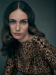 Close-up portrait of a woman with wavy brunette hair wearing a high-neck leopard-print top with shoulder buttons.