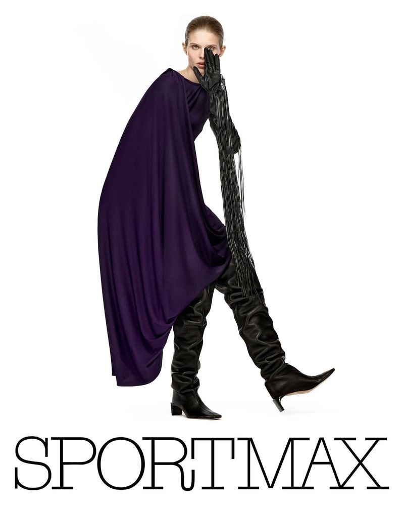 Fashion-forward portrait of a slender adult female wearing a long purple dress, leather gloves with long fringe, and thigh-high leather boots.