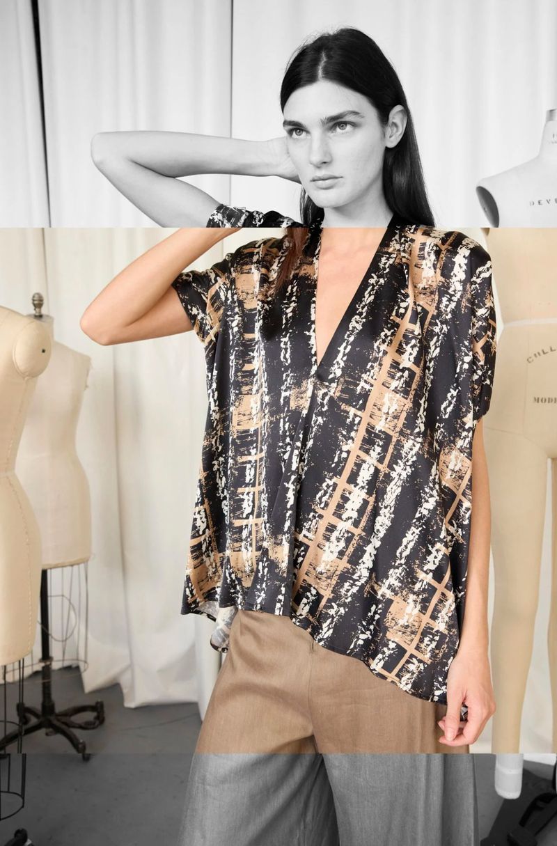Fashion shot of a woman in a printed top and tan pants, posing in a studio, with neutral backdrop and dressmaker's mannequins visible.