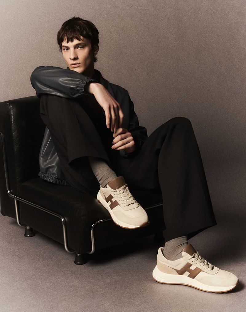 Adult male model sits on a black chair wearing a black jacket, dark pants, and light trainers in a studio setting with a gray background.