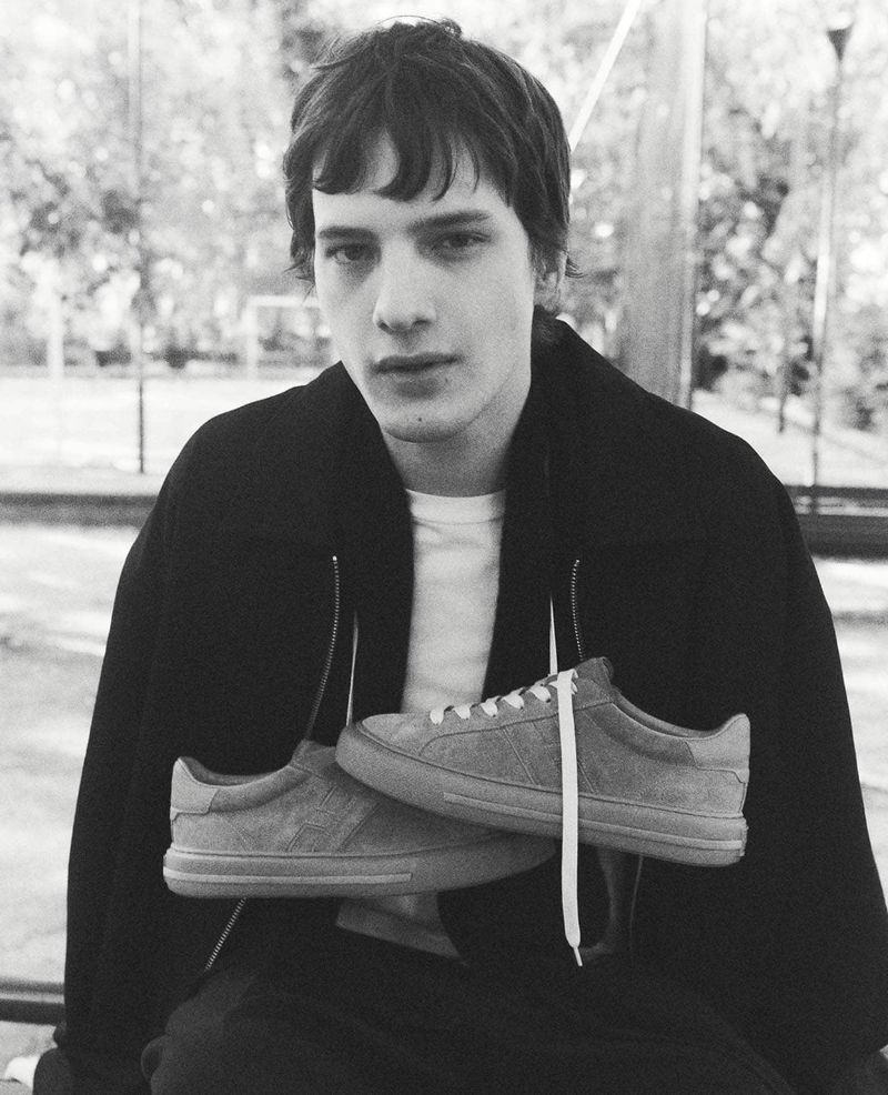 Black and white portrait of a young adult male in a jacket, seated outdoors, holding a pair of casual sneakers with white laces.