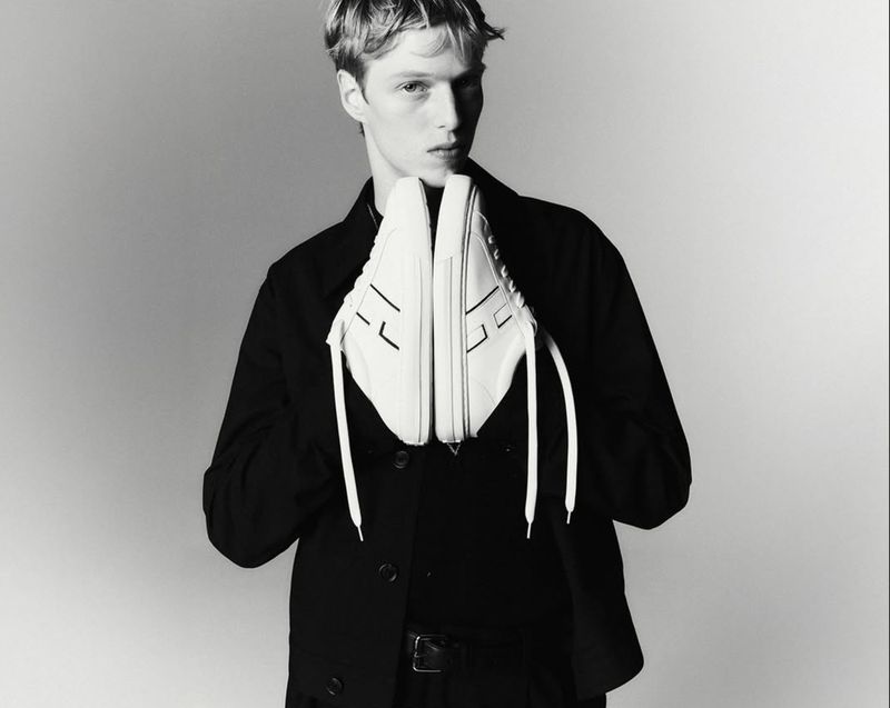 Monochrome studio shot of a young male adult holding a pair of white sneakers with black details, wearing a black jacket.