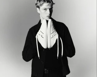 Young male model in a black jacket holding up a pair of white sneakers against a light grey seamless studio background.