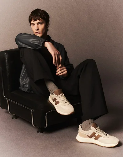 Model seated in a black leather armchair wearing dark jacket and trousers with beige sneakers, for Hogan F/W 2025.