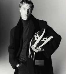Monochrome portrait of a young male model holding sneakers, wearing a dark coat and turtleneck against a plain studio background.
