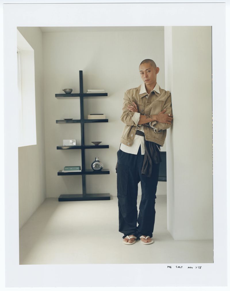 An adult male model in a casual outfit leans against a column indoors, next to a black bookshelf in a light-filled minimalist space.