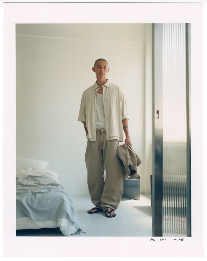 Male fashion model posing indoors in casual linen shirt, pants, and sandals; soft lighting, with a bed and window in background.