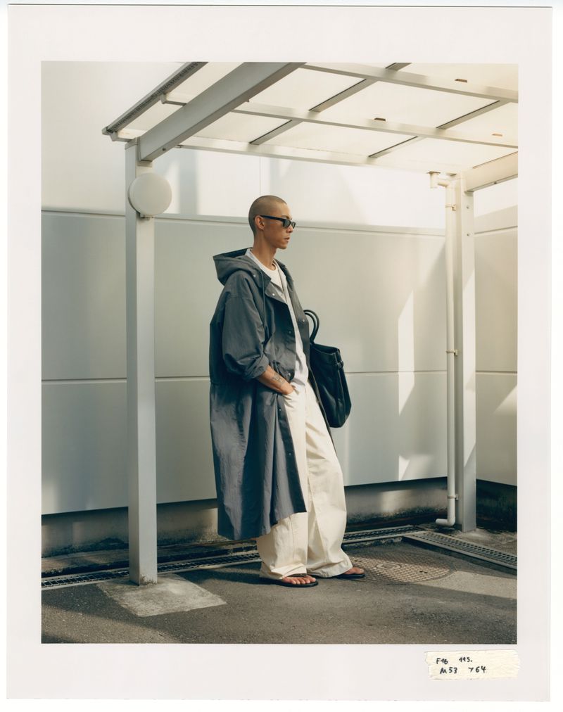 A bald adult in a hooded overcoat, sunglasses, and sandals poses on an urban sidewalk near a building with white walls and a metal awning.
