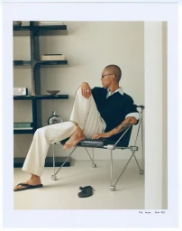 Model with shaved head wearing dark polo and white trousers seated in a modern metal chair against neutral shelving