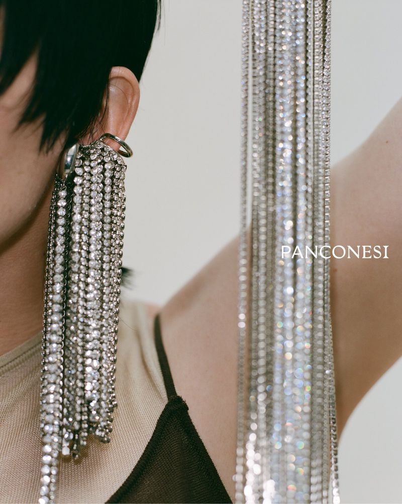 Close-up studio shot of an adult female model with short dark hair wearing a tan top with silver rhinestone fringe earrings.