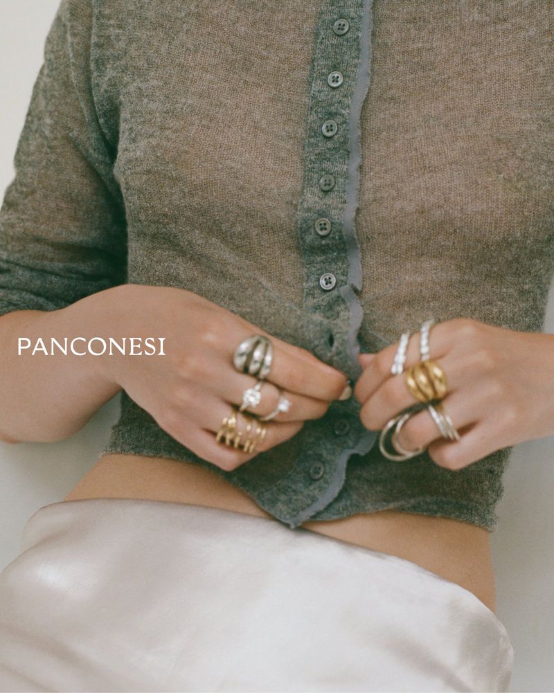 Close-up of a model wearing stacked silver and gold sculptural rings while holding a sheer grey knit cardigan over a silk skirt.