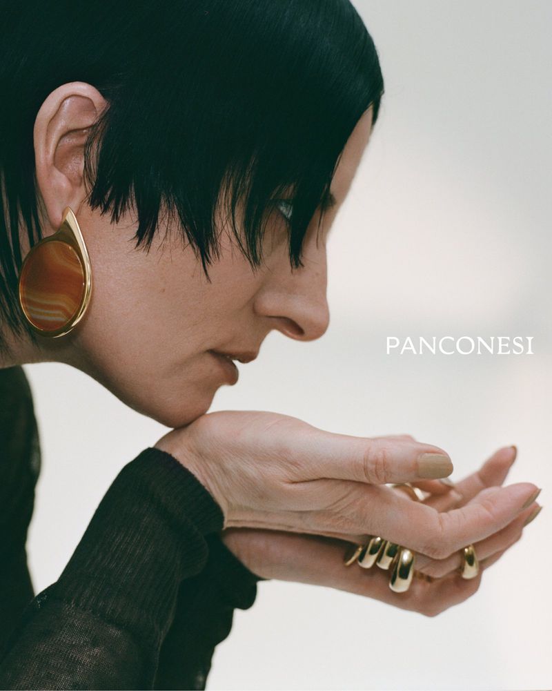 Model in profile wearing a statement gold drop earring and chunky gold rings against a clean light background.
