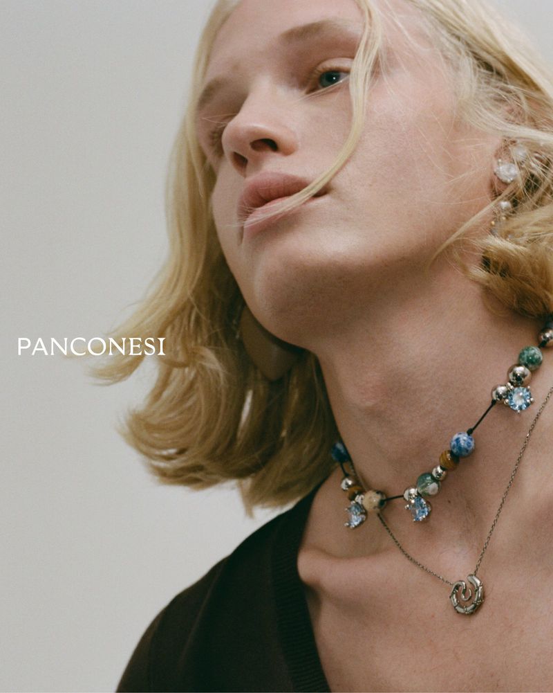Close-up of a young adult with blonde hair wearing beaded necklace and silver pendant against a neutral backdrop in soft lighting.