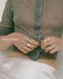 Close-up of a model wearing stacked silver and gold sculptural rings while holding a sheer grey knit cardigan over a silk skirt.