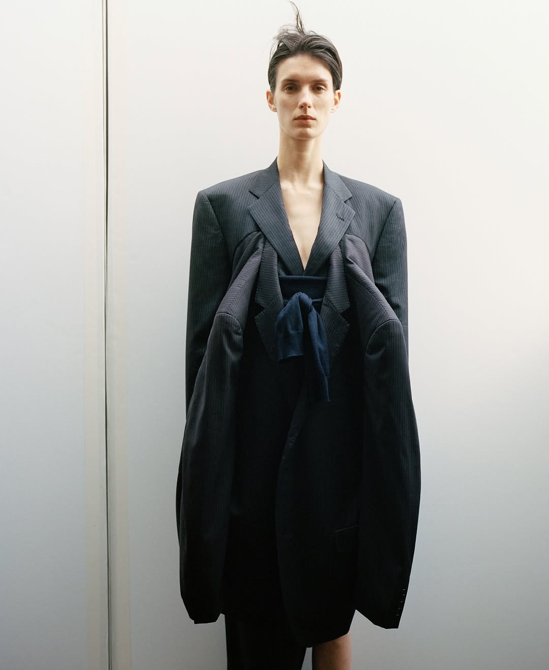 Fashion portrait of a slender person in a pinstripe suit holding a larger jacket, against a minimalist off-white wall backdrop.