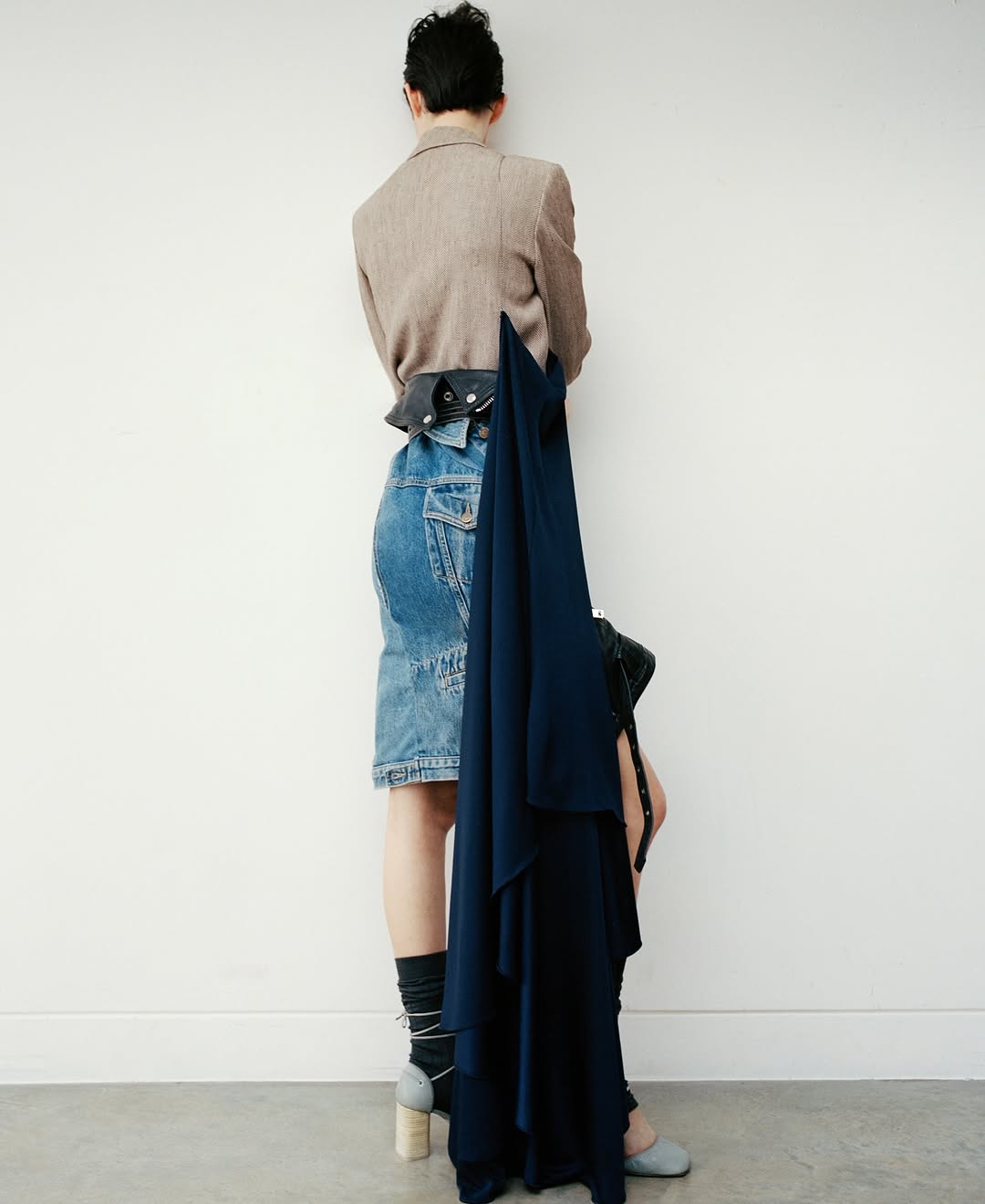 Back view of a woman in a beige blazer, denim skirt, and grey heels against a white wall, holding a dark blue draped dress.
