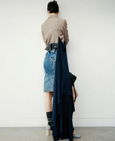 Back view of a woman in a beige blazer, denim skirt, and grey heels against a white wall, holding a dark blue draped dress.