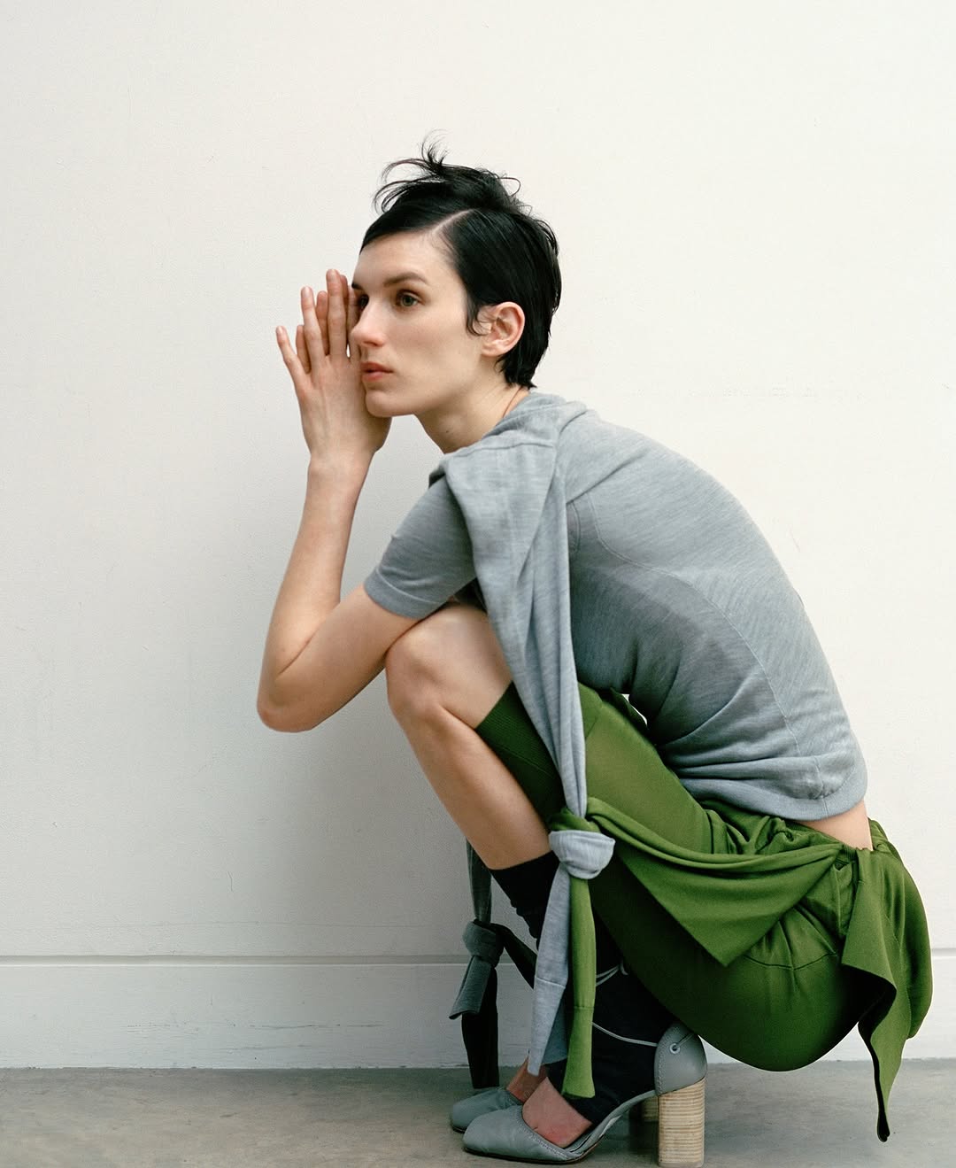 Woman in grey top and green skirt sits in studio with hands together in thoughtful pose, neutral background, professional fashion shoot.