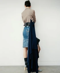 Model standing with back turned wearing a tan blazer and denim skirt against a white studio background.