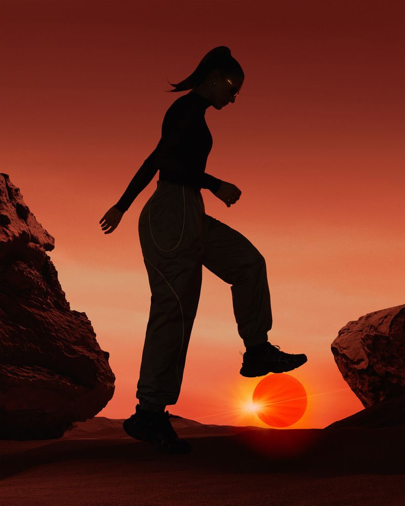 Silhouette of a female figure stepping from one rock formation to another against an orange sky with the sun appearing between rocks.