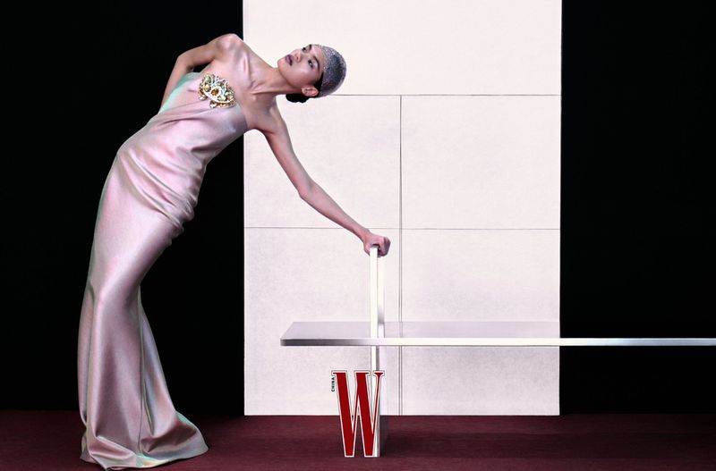 Elegant woman in a pink gown poses with a modern table against a black and white geometric backdrop in a fashion editorial shot.
