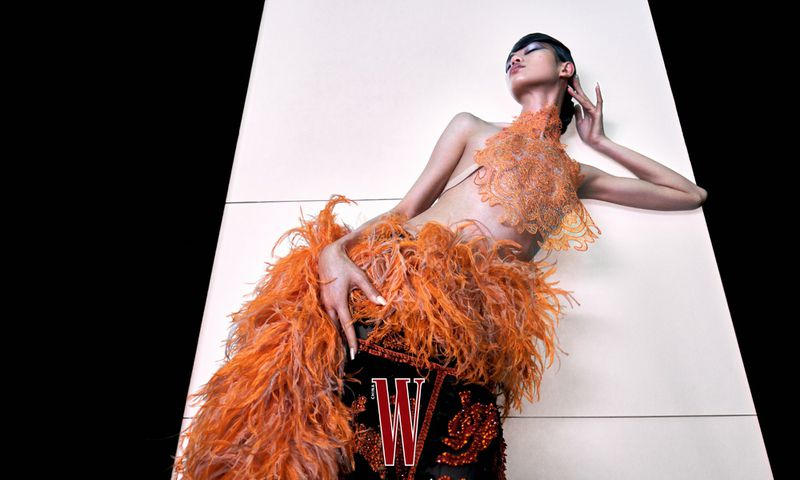 Fashion shot of a model in orange feathered garment, laying against a white panel with black background. 'W' magazine logo present.