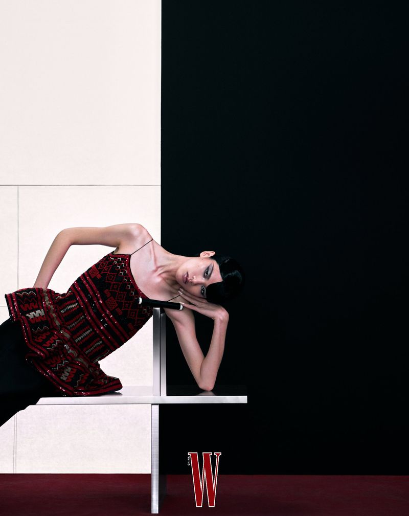 Fashion editorial shot of a woman in patterned top and dark pants lying on a white ledge against a black and white background.