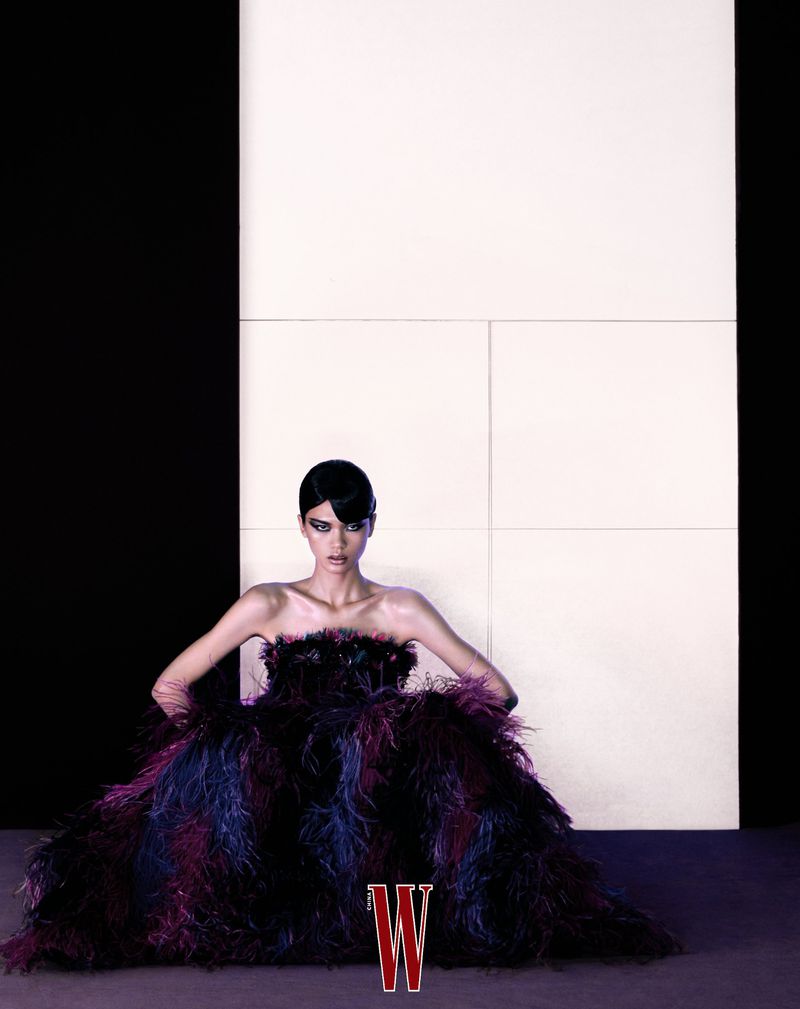 Elegant adult female model in strapless feathered gown poses indoors against black and white panels in a studio setting for fashion editorial.