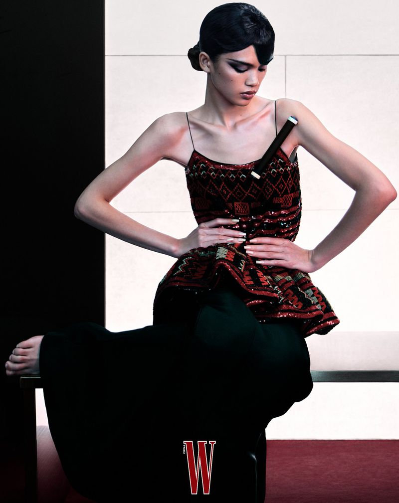 Fashion photo of a slender woman posing in a patterned top and dark wide-leg pants. She is seated on a platform with a minimalist backdrop.