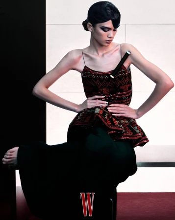 Fashion photo of a slender woman posing in a patterned top and dark wide-leg pants. She is seated on a platform with a minimalist backdrop.