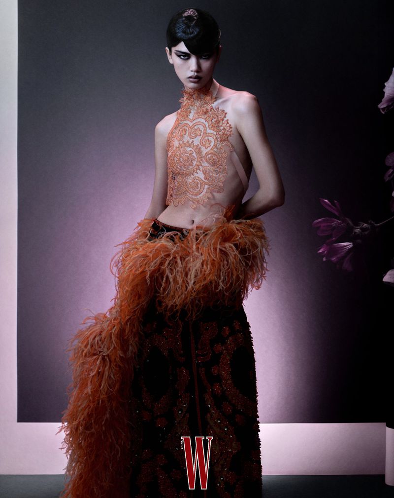 A fashion model poses in a dress with a feathery boa. Editorial fashion shot against a gradient grey-purple background in a photo studio.