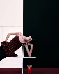 Model Qun Ye leans against a geometric metal bench wearing an embellished burgundy peplum top against a split black and white studio wall.