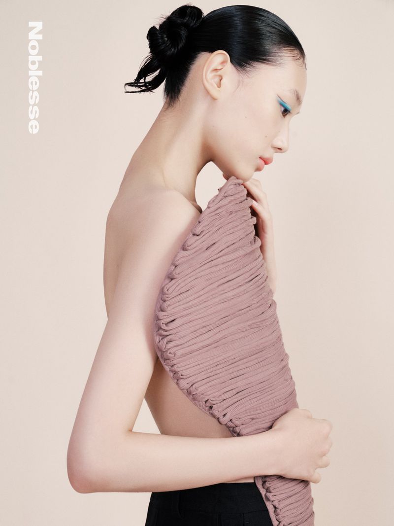 Side profile of an Asian woman with a bun hairstyle, wearing blue eyeshadow, holding a textured mauve fabric against a beige backdrop.