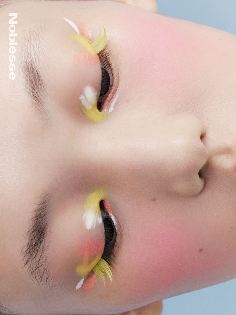 Close-up shot of a young adult's face featuring artistic yellow and white feathered false eyelashes and pink blush makeup.