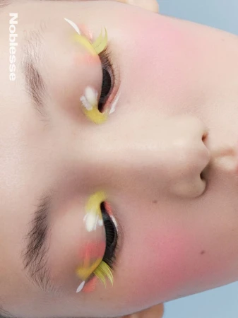 Close-up shot of a young adult's face featuring artistic yellow and white feathered false eyelashes and pink blush makeup.