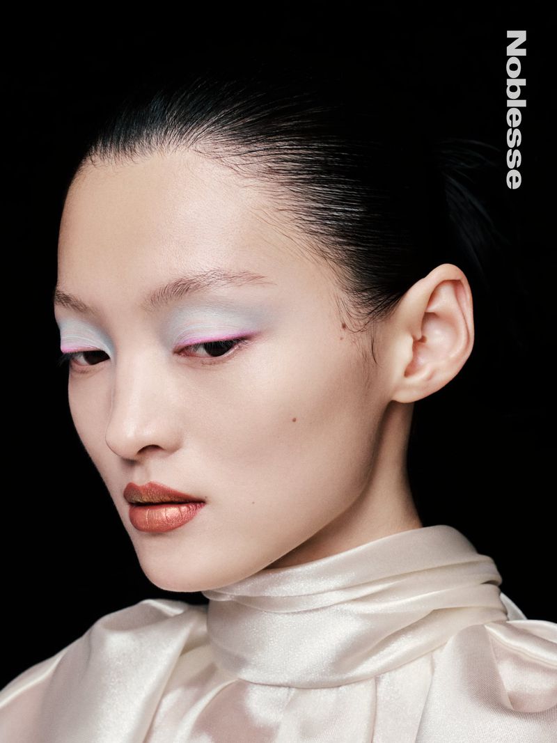 Close-up beauty shot of a young Asian woman with glossy bronze lipstick and artistic white eyeshadow, against a stark black backdrop.