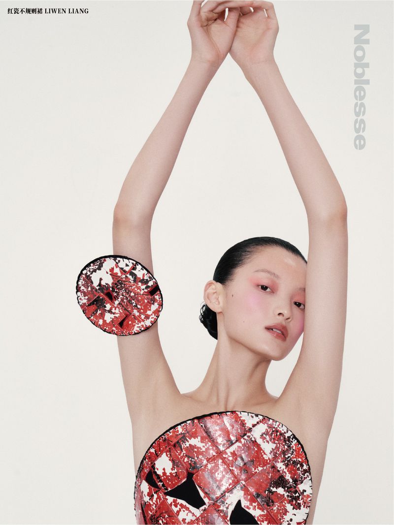 Editorial fashion shot of a bare-chested Asian model with arms raised wearing round red-and-white avant-garde discs against a white backdrop.