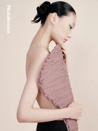 Model with dark hair in a bun wearing a textured mauve garment and black trousers against a neutral background.
