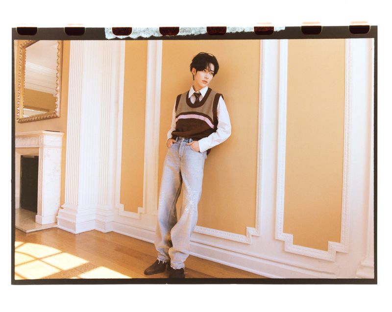 Portrait of an East Asian male in a sweater vest and jeans posing in a neutral-toned interior with ornate wall trim and sunlight.